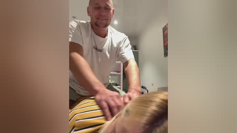 Media: Video of a bald, muscular man in a white t-shirt giving a shoulder massage to a blonde man in a yellow and white striped shirt. Background shows a blurred room with a wall and a framed picture.