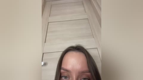 Video of a young Caucasian woman with straight brown hair and light skin, looking down from an elevated angle. The background features beige wooden stairs and a handrail, creating a minimalistic, modern indoor setting.