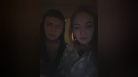 Video of two young women with fair skin and light brown hair, wearing dark clothing, standing close together in a dimly lit room with a blurred background.