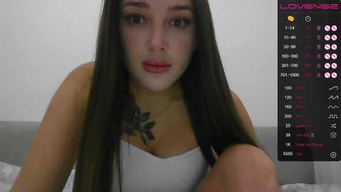Video of a young woman with long dark hair, fair skin, and a floral tattoo on her chest. She wears a white tank top, with a video feed showing social media stats in the background.