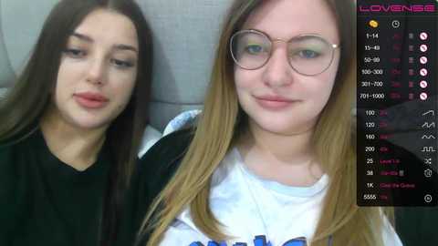 Video of two young women, one with long brown hair, the other with long blonde hair, both wearing glasses, smiling, in a cozy indoor setting with a gray couch.