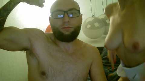 Media: Video of a shirtless, muscular man with glasses and a beard flexing his arm in front of a mirror. Behind him, a woman's bare chest is visible.