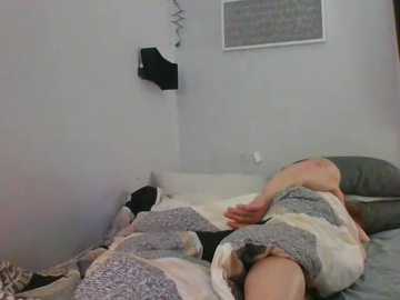 Video of a person lying in bed, partially covered by a patterned blanket, with a black shirt and white socks visible. The room has light gray walls and a small window with a white blind.
