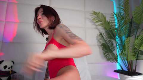 Video of a slender, tattooed woman with short dark hair in a red one-piece swimsuit, dancing in a modern room with white walls, green plants, and pink and blue neon lights.