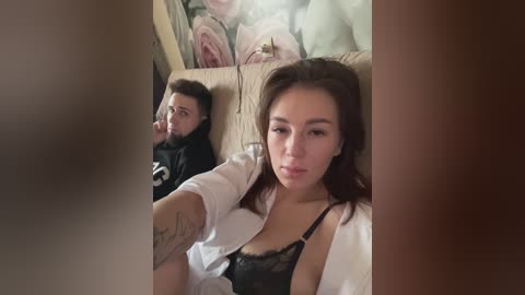 Media: Video of a young woman with light skin and brown hair, wearing a black lace bra and white robe, sitting on a beige couch. A man with a beard and dark hair is visible in the background, also sitting on the couch.