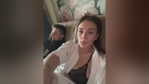Media: A video captures a woman with long dark hair, wearing a black lace bra under an open white shirt, sitting on a beige couch in a dimly lit room with floral wallpaper.
