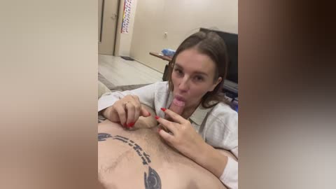Media: Video of a white woman with long brown hair, wearing a white robe, performing oral sex on a man with a tattooed penis in a clinical room.