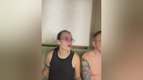 Video of a topless tattooed man with glasses and a woman with pink glasses and black tank top, both seated, partially obscured by a blurred foreground.