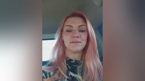 Video of a young Caucasian woman with straight, long, pink hair, wearing a black, patterned top, looking down and slightly to the left. Background is blurred, showing a car interior.