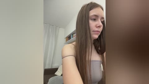 A video of a young Caucasian woman with long brown hair and a neutral expression, wearing a light grey spaghetti-strap top, standing in a simple, white-walled room with a bookshelf and curtains.
