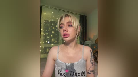 Media: Video of a young, fair-skinned, blonde-haired woman with a slender physique wearing a grey tank top with \"Yes Daddy\" text. She has a tattoo on her left arm. Background features a dimly lit room with a fairy light string and a window.