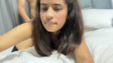 A video of a young woman with medium brown skin and long, straight brown hair, lying on a white bed, wearing a white shirt. The background features light gray curtains and a pillow.