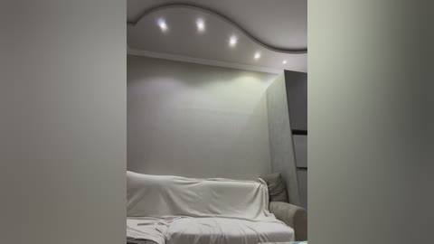 Video of a minimalist living room with a white sectional sofa draped in a white blanket. The walls are plain and light-colored, with recessed ceiling lights. The room's decor is modern and clean, emphasizing simplicity.