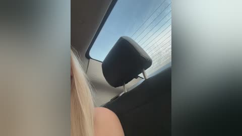 Video of a car interior, focusing on a black leather headrest with a white metal frame, positioned against a clear, blue sky visible through a large window. The car seat is beige and the side panels are grey.