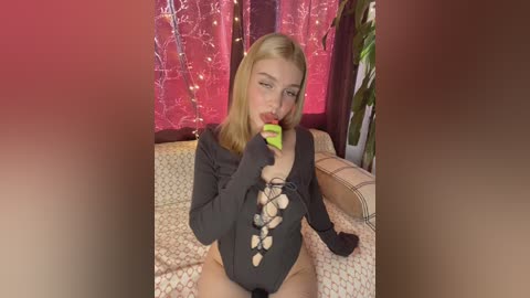 Media: Video of a blonde woman with fair skin, wearing a black lace bodysuit, biting a green fruit, seated on a patterned couch.