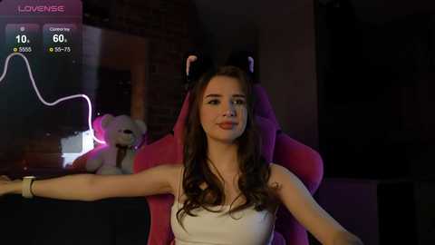 Media: Video of a young woman with long brown hair, wearing a white dress and black cat ears, sitting in a pink gaming chair. Background includes a dimly lit room with a teddy bear, a brick wall, and a glowing digital screen.