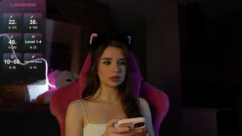 Media: A video of a young woman with light skin and long brown hair, wearing a white camisole, playing a video game on a pink gaming chair.