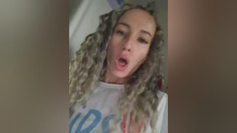 Video of a young, light-skinned woman with curly, grey hair and a white T-shirt, captured in a blurry, close-up selfie. She's sticking her tongue out playfully.