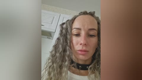 A video of a woman with long, wavy, gray hair and a black spiked collar, standing in a kitchen with white cabinets and a beige countertop.