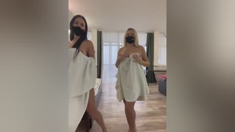 Media: Video of two women in a modern, bright room. Both are wrapped in white towels, one wearing a black face mask, standing near a white sofa.