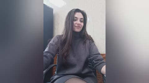 Media: A video of a young woman with long dark hair, wearing a dark sweater, seated in a chair against a beige wall, with blurred objects in the foreground.