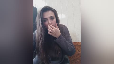 Media: Video of a young woman with long, straight brown hair, wearing a gray sweater, and a confused expression, standing in a room with white paneled walls and a wooden panel on the right.