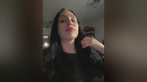 Media: Video of a young woman with long black hair, wearing a dark top, standing in a dimly lit room with a blurred background.