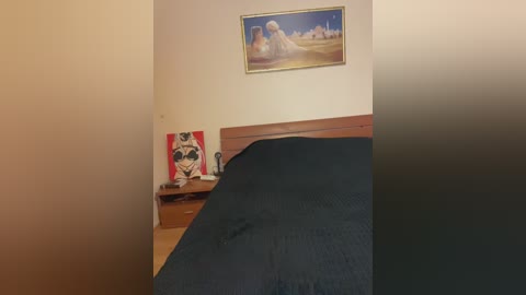 Video of a minimalist bedroom with a beige wall, wooden headboard, framed painting of a white angel, and a black comforter.