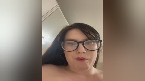 Media: Video of a light-skinned woman with long brown hair, wearing black-rimmed glasses, red lipstick, and hoop earrings. She has a neutral expression, and the background is blurred with a beige wall and ceiling.