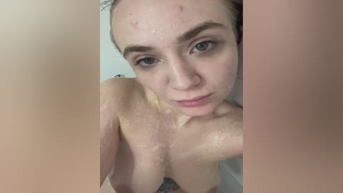 A video of a topless woman with fair skin, wet hair, and light eyes, gazing directly into the camera. She has medium-sized breasts and a neutral expression. The background is blurry.
