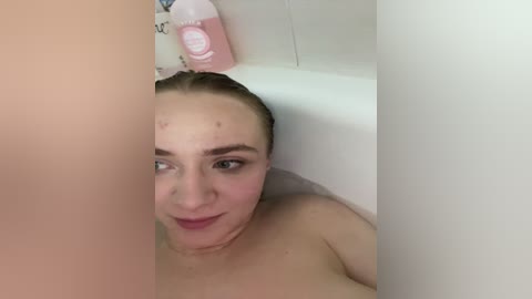Video of a young woman with light skin and blonde hair, partially submerged in a white bathtub, smiling slightly. A pink bottle of shampoo is visible in the background.