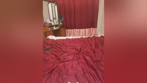Video of a messy bedroom with a red bedspread, a wooden dresser, a mirror, and a maroon curtain.