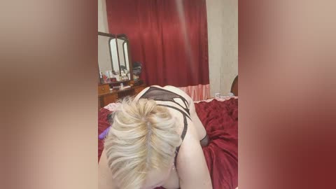 Video of a blonde woman in black lingerie on a bed, facing away from the camera, with red curtains and a dresser in the background.