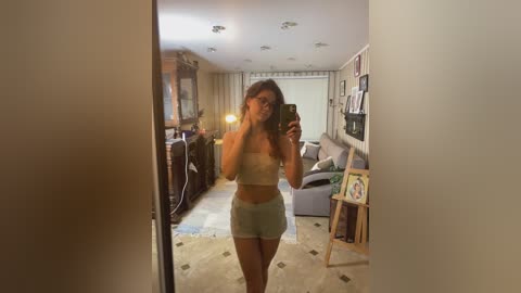A video of a young woman with light skin and brown hair, wearing a white bandeau top and light gray shorts, taking a mirror selfie in a cozy, warmly lit living room with beige carpeting and bookshelves.