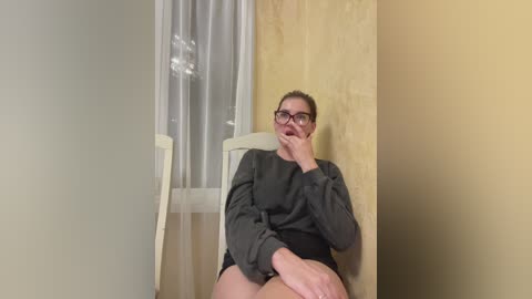 Media: Video of a white woman with glasses, medium build, wearing a gray sweater and black shorts, sitting on a white chair in a corner with a beige wall and a light wooden panel.