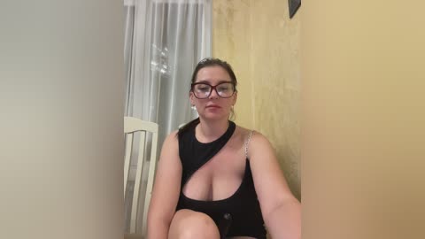 Media: Video of a plus-sized woman with glasses, black one-shoulder top, and hoop earrings, sitting on a white chair in a beige room with a yellow wall.