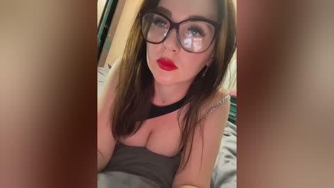 Media: Video of a Caucasian woman with long brown hair, wearing large-framed glasses, red lipstick, and a black choker, lying on a bed with gray sheets, viewed from a low angle.