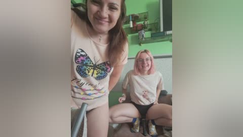Video of two women, one with fair skin and brown hair, wearing a tie-dye shirt, the other with light skin and pink hair, wearing a white shirt and black underwear, sitting on a couch.