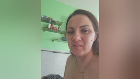 Video of a middle-aged woman with light skin and brown hair, wearing a necklace, standing in a green-painted room with a grey towel and shelves holding toiletries.