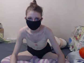 Video of a slender, fair-skinned woman with light brown hair in a bun, wearing a black face mask and black shorts, sitting on a bed with a floral-patterned pillow, in a cluttered room.