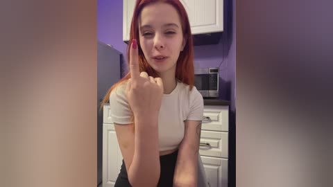 Video of a young woman with long, straight red hair, fair skin, and a petite frame. She is in a modern kitchen, wearing a white crop top and black pants, making a fist with her right hand.