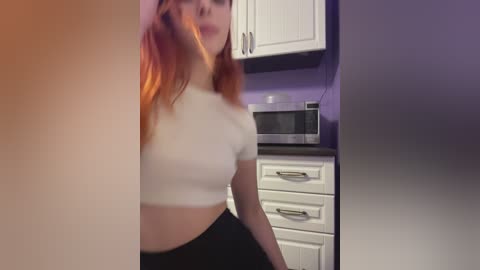 Video of a young woman with long, vibrant orange hair, wearing a tight, white crop top and high-waisted black skirt, standing in a modern kitchen with white cabinets and a microwave.