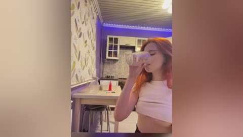 Video of a young woman with long orange hair, wearing a white crop top, drinking from a glass in a modern kitchen with light wood countertops and floral-patterned wallpaper.