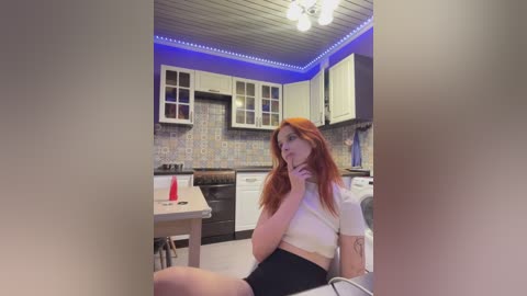 Video of a young woman with long red hair, wearing a white crop top and black shorts, sitting in a modern kitchen with blue LED strip lights, white cabinets, and a tiled backsplash.