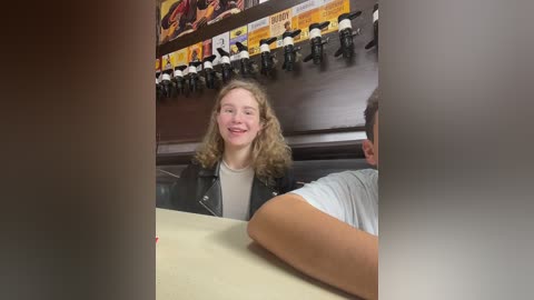 Media: Video of a smiling young woman with curly blonde hair in a black jacket, seated at a table, with a man's back in a white shirt partially visible. Background includes a wall with racing-themed memorabilia.