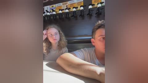 Media: Video of a young woman with curly hair and a young man with short hair, both seated in a dimly lit room with a wall-mounted collection of toy soldiers.