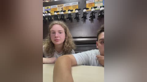 Media: Video of two young women with long hair, sitting at a table in a dimly lit room. One woman, with curly hair, wears a gray shirt, while the other, with straight hair, dons a white top. Background shows a shelf with multiple microphones.