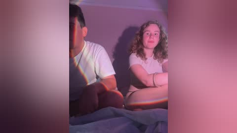 Media: Video of a young couple sitting on a bed, with a rainbow prism effect cast on them. The man has short dark hair, wearing a white T-shirt. The woman has curly blonde hair and a white top. They have serious expressions.
