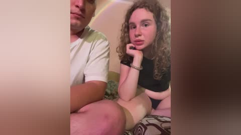 Media: Video of a man and woman, both with light skin, sitting on a bed with floral sheets. The woman, with curly hair, wears a black crop top, and the man, in a white t-shirt, has a neutral expression.