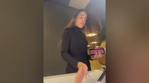 Media: Video of a young woman with long brown hair in a black turtleneck, holding a purple drink bottle in a modern, dimly lit bathroom with a gold-colored wall and white countertop.